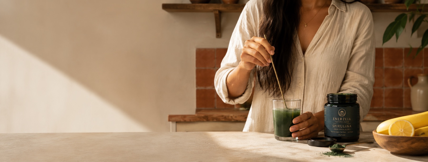 A woman at a sunlit kitchen counter stirring a glass of deep green spirulina drink with a long spoon, an open Enerzeia Spirulina jar and a bowl of lemons and bananas beside her.