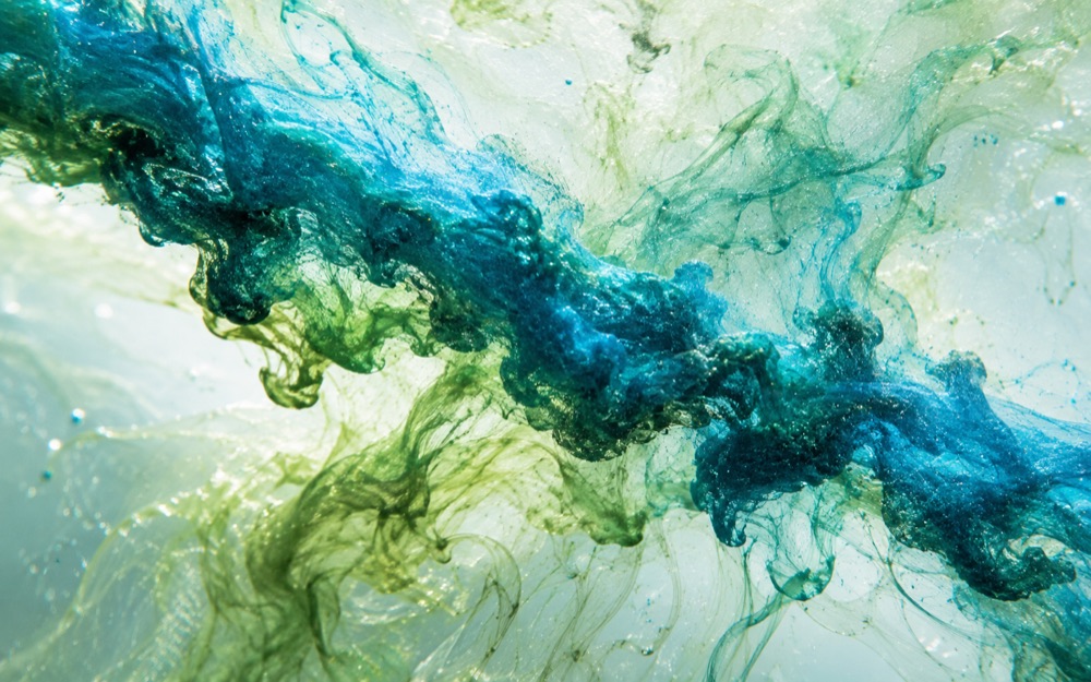 A macro photograph of blue and green pigment dispersing through water in fine billowing threads against a pale background.
