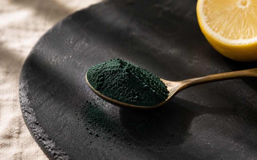 A brass spoon heaped with dark green spirulina powder resting on a round slate plate in low sunlight, a cut lemon half beside it.